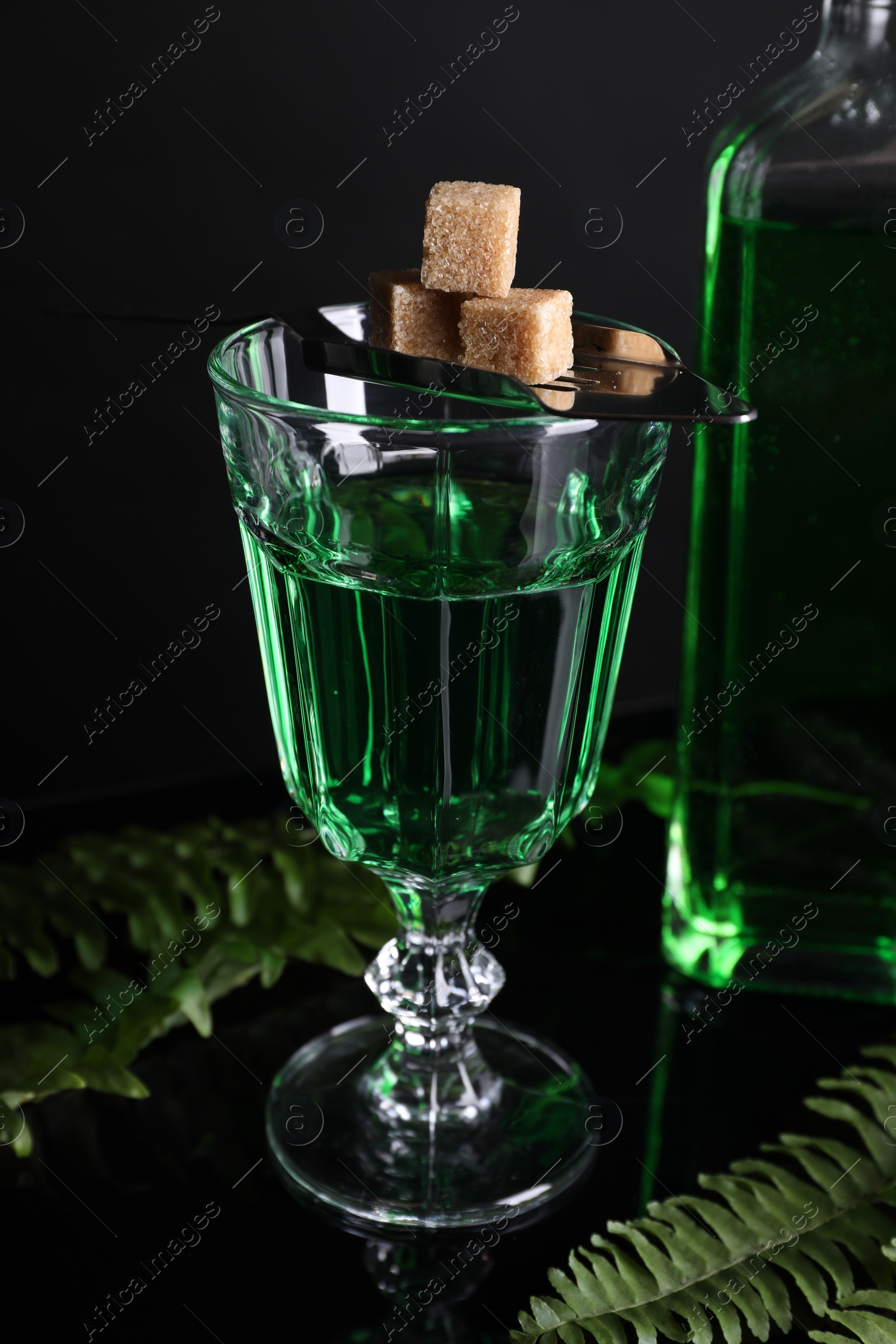 Photo of Absinthe in glass, brown sugar, spoon and green leaves on mirror table. Alcoholic drink