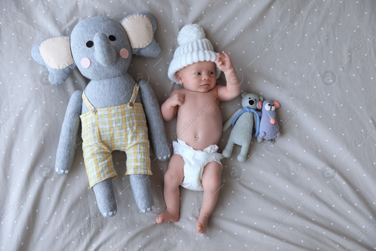 Photo of Cute newborn baby in white knitted hat with toys on bed, top view