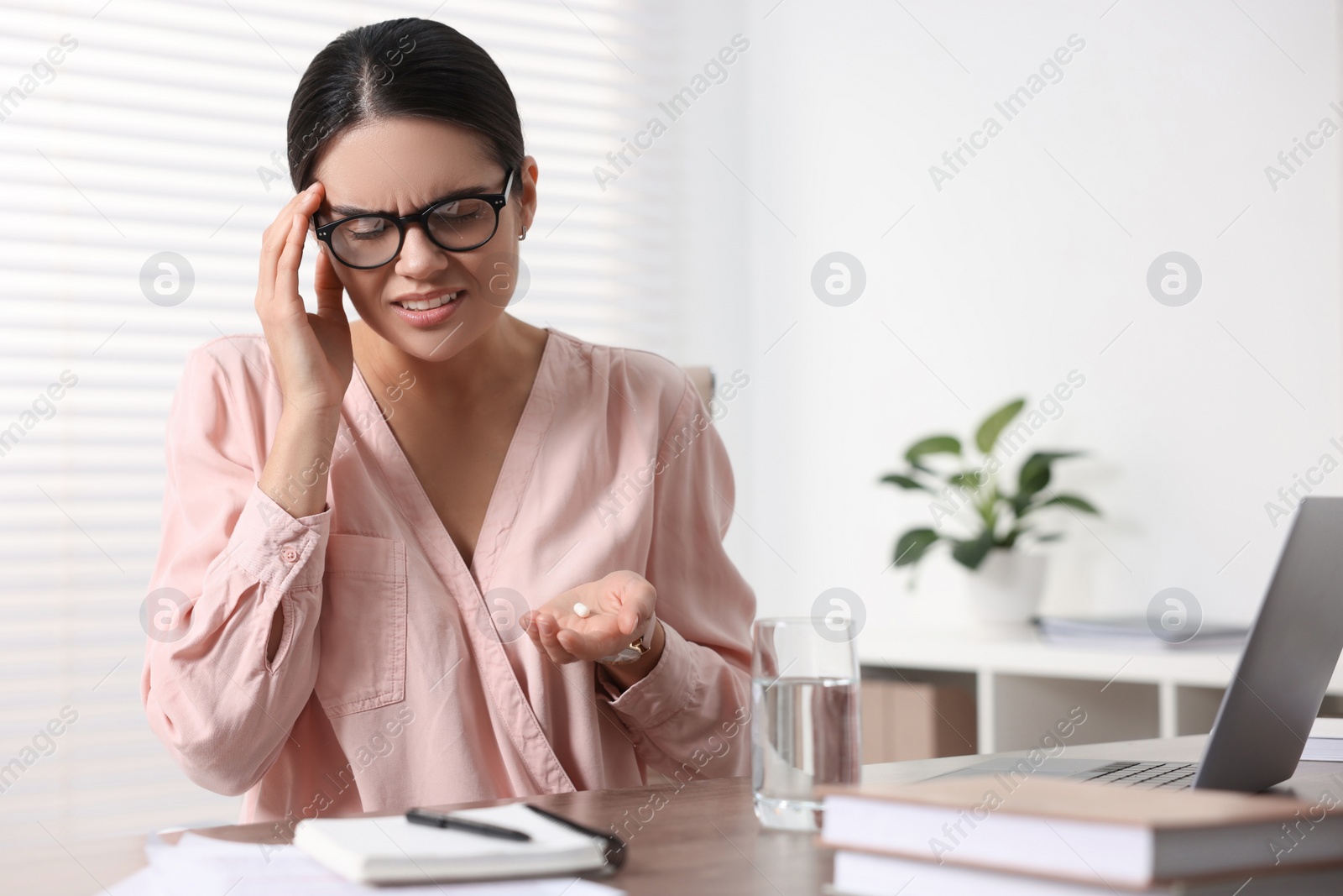 Photo of Young woman with pill and glass of water suffering from headache at wooden table in office