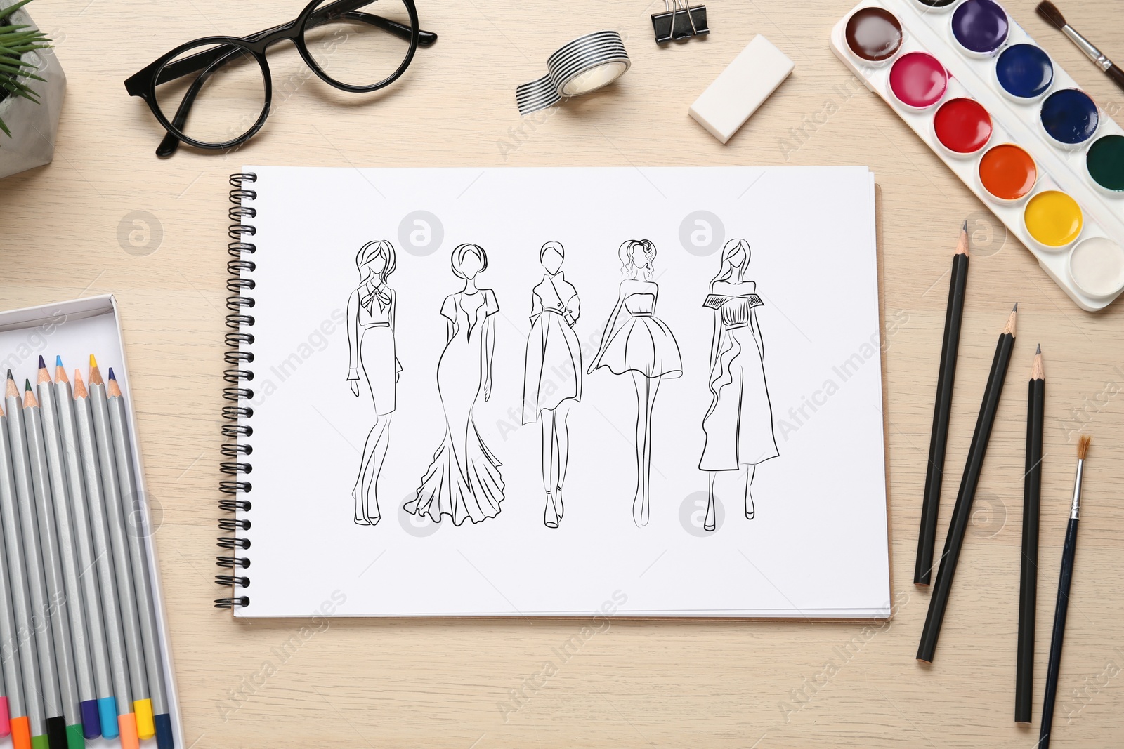 Image of Sketches of different clothes in pad on wooden table. Fashion designer's desk with stationery, flat lay