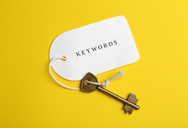Photo of Metal key and tag wIth word KEYWORDS on yellow background, top view