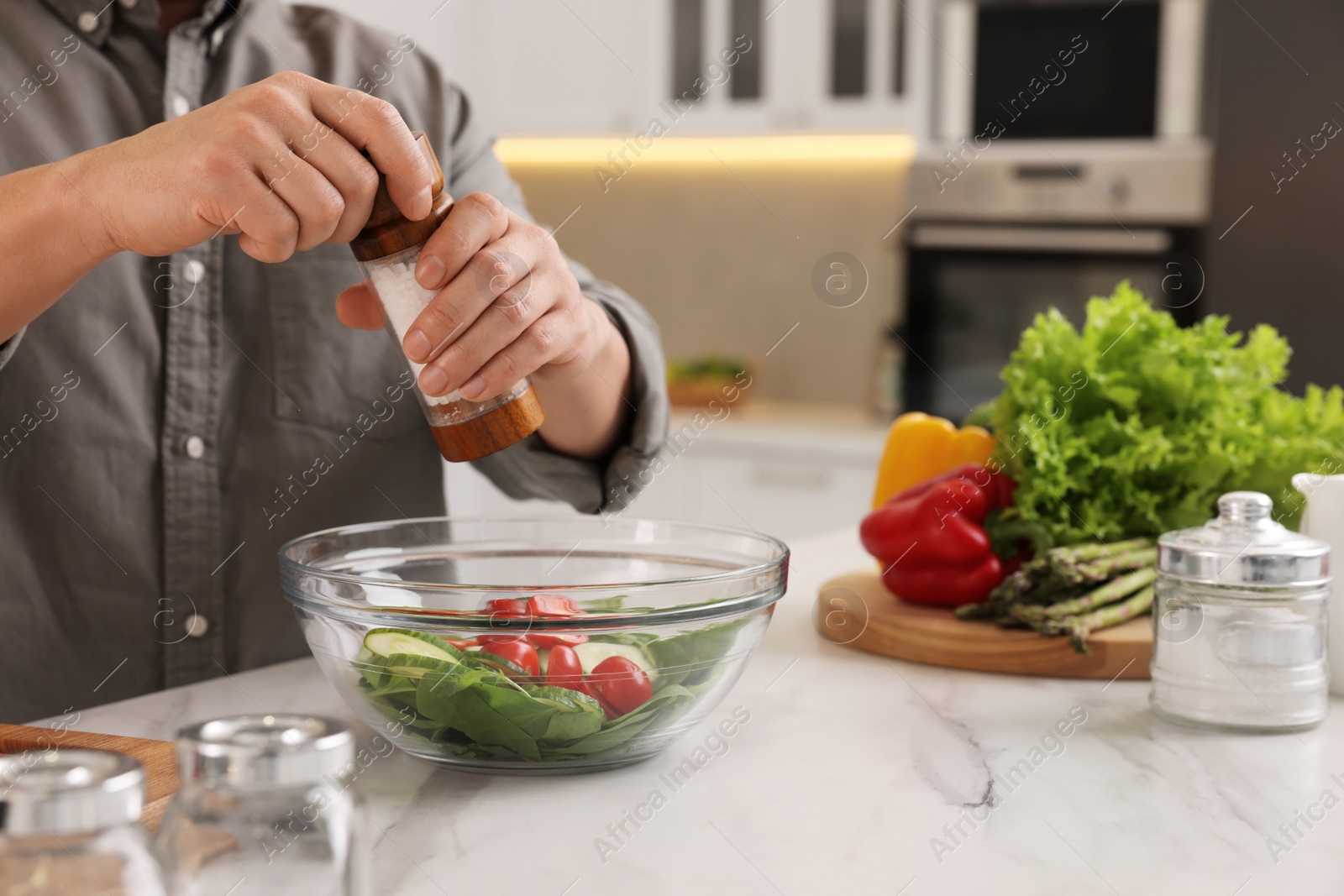 Photo of Cooking process. Man adding salt into bowl of salad at white marble countertop kitchen, closeup
