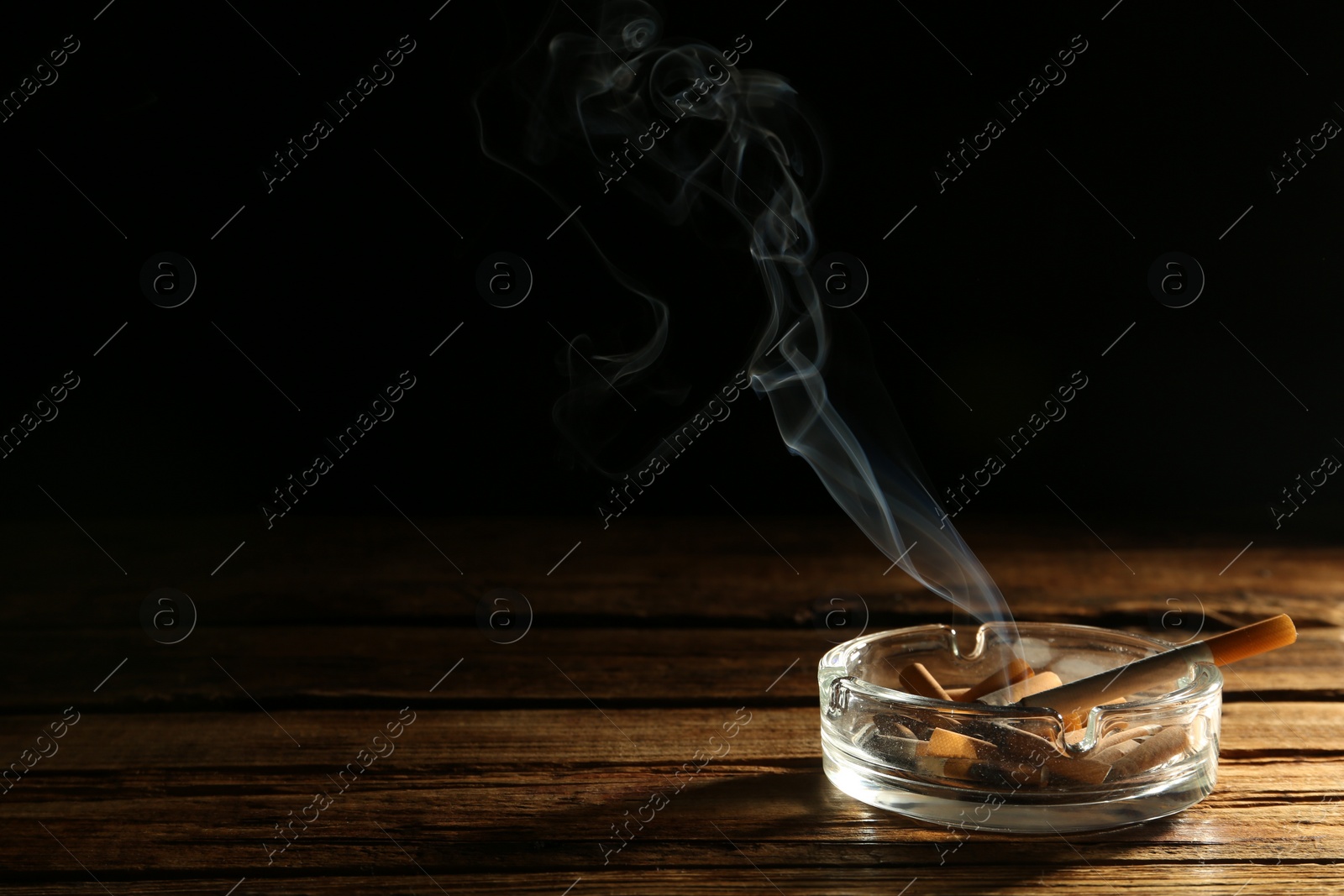 Photo of Smoldering cigarette in glass ashtray on wooden table against black background. Space for text