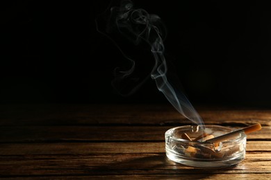 Photo of Smoldering cigarette in glass ashtray on wooden table against black background. Space for text