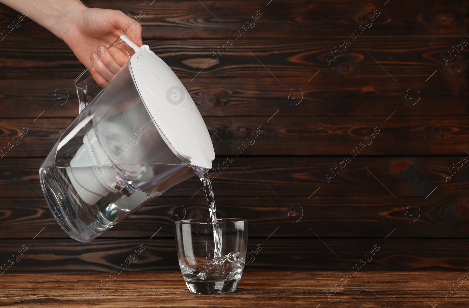 Photo of Woman pouring purified water from filter jug into glass on wooden table, closeup. Space for text