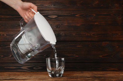 Woman pouring purified water from filter jug into glass on wooden table, closeup. Space for text