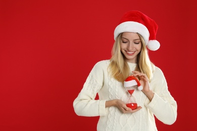 Photo of Woman in Santa hat with hourglass on red background, space for text. Christmas countdown