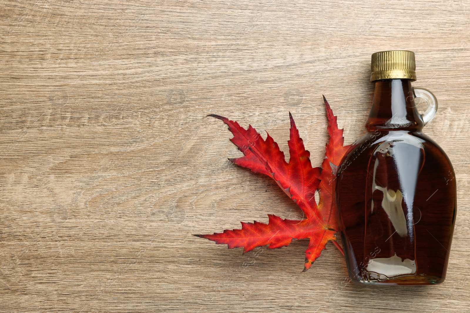 Photo of Glass bottle of tasty maple syrup and dry leaf on wooden table, flat lay. Space for text