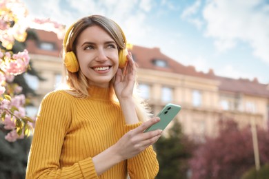 Young woman with smartphone and headphones listening to music outdoors on sunny day, space for text