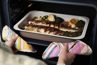 Photo of Woman taking out baking tray with sea bass fish and vegetables from oven, closeup