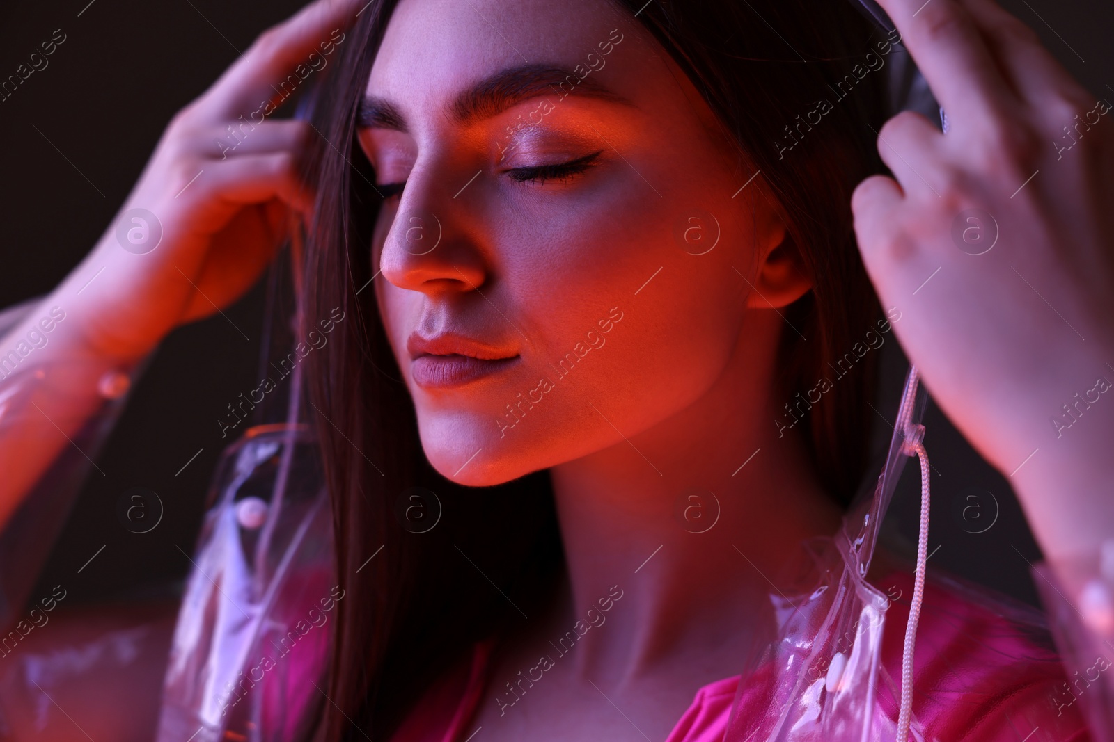 Photo of Fashionable portrait of beautiful woman wearing transparent coat on dark background in neon lights, closeup