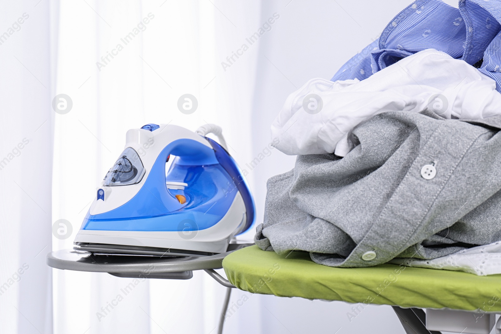 Photo of Modern electric iron and heap of clothes on board indoors. Laundry day