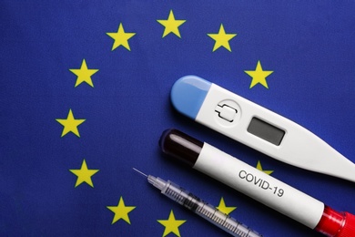 Test tube with blood sample, thermometer and syringe on European Union flag background, flat lay. Coronavirus outbreak