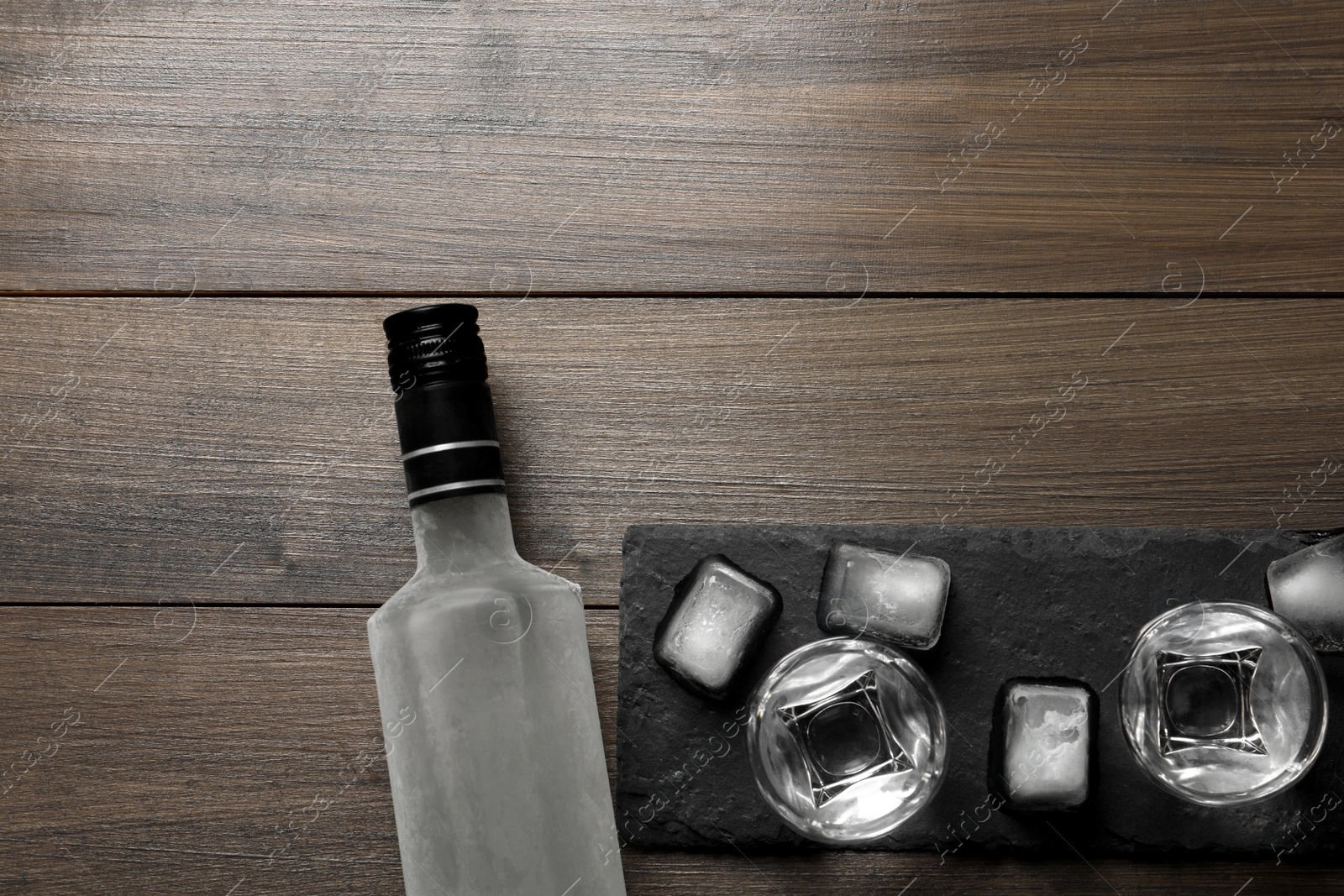 Photo of Bottle of vodka, shot glasses and ice cubes on wooden table, flat lay. Space for text