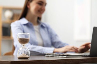 Photo of Hourglass with flowing sand on desk. Woman using laptop indoors, selective focus