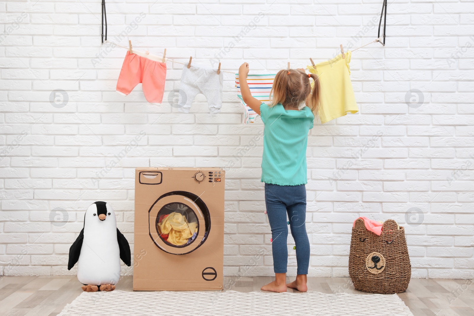 Photo of Little girl hanging clean laundry near toy cardboard washing machine indoors