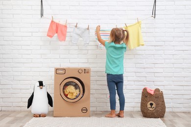 Little girl hanging clean laundry near toy cardboard washing machine indoors