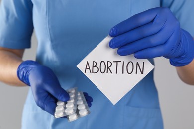 Photo of Doctor holding pills and paper card with word ABORTION on light grey background, closeup
