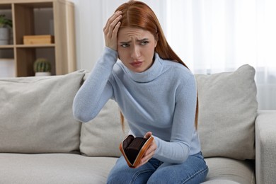 Upset woman with empty wallet on sofa indoors