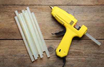 Photo of Yellow glue gun and sticks on wooden table, flat lay