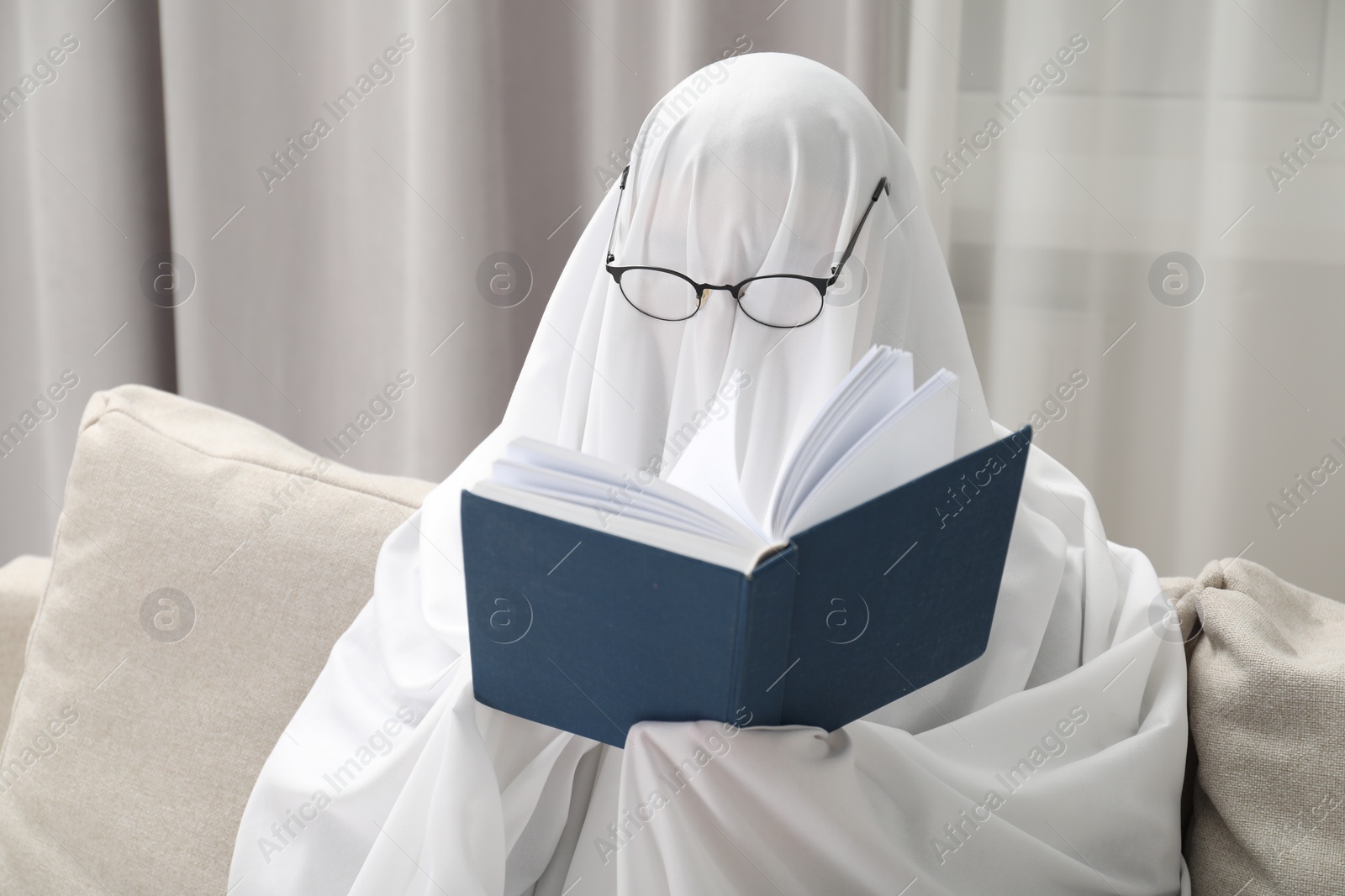 Photo of Creepy ghost. Person covered with white sheet reading book on sofa at home