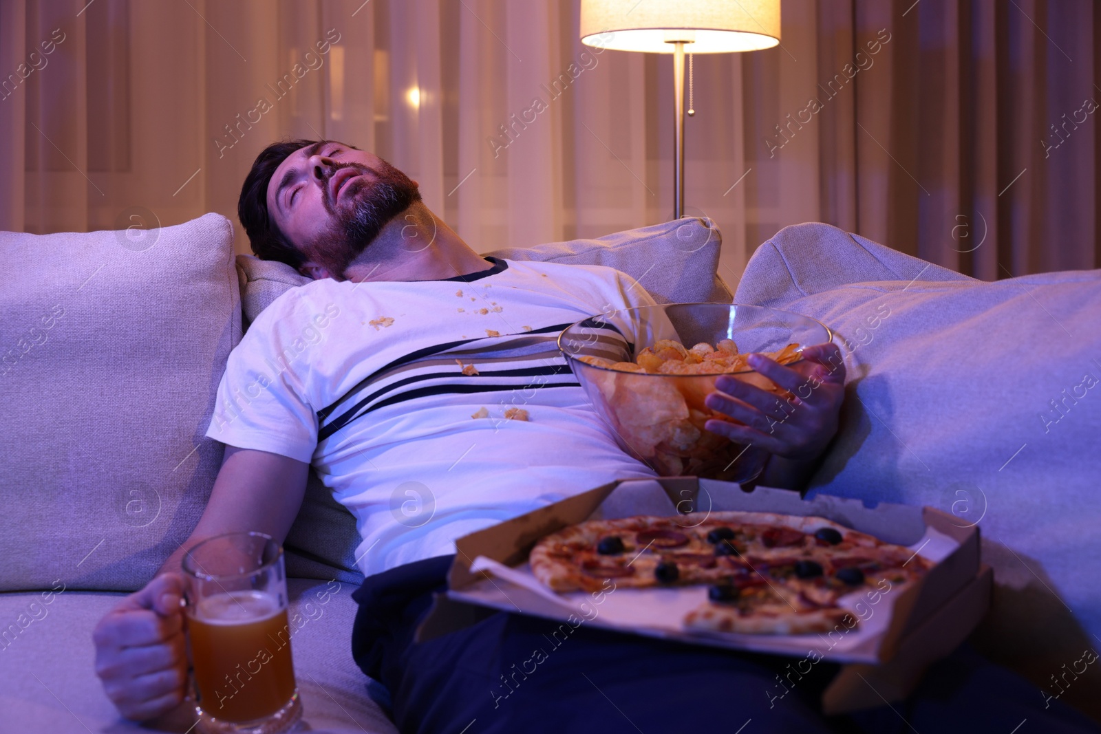 Photo of Man with chips, pizza and glass of beer sleeping on sofa at night. Bad habit