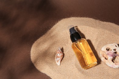 Photo of Bottle of serum, seashell and stone on sand against brown background. Space for text