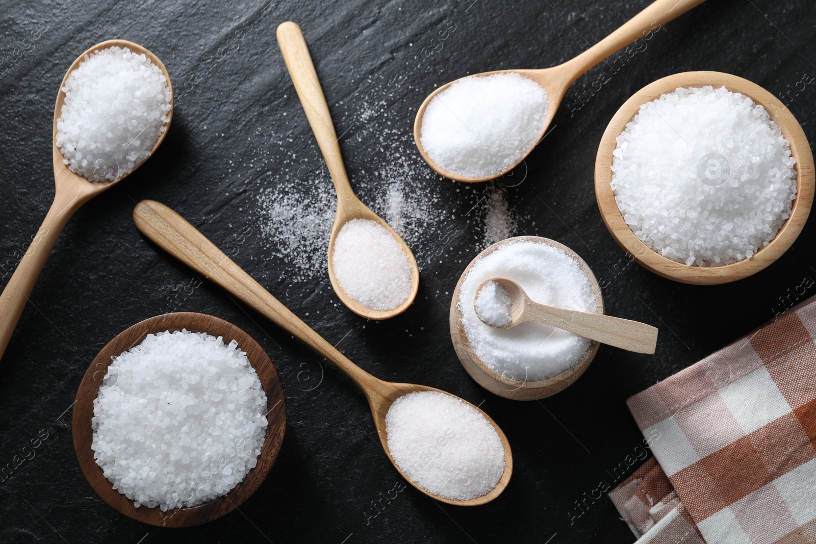 Photo of Organic white salt in bowls and spoons on black table, flat lay