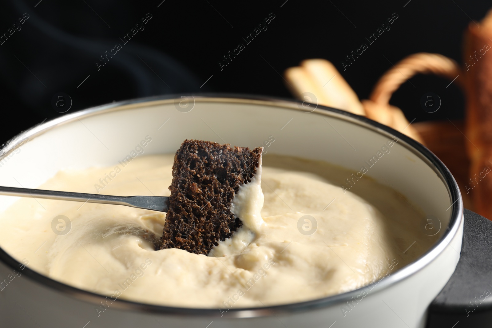 Photo of Dipping piece of bread into fondue pot with melted cheese on black background, closeup