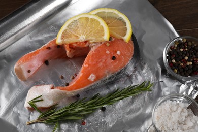 Photo of Aluminum foil with raw fish, lemon slices, rosemary and spices on wooden table, closeup. Baking salmon