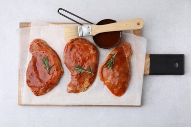 Board with raw marinated meat, rosemary and basting brush on light table, top view