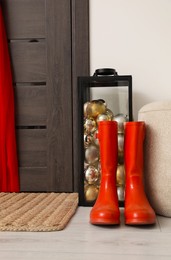 Container with baubles, ottoman and red rubber boots near wooden door in room. Christmas decoration