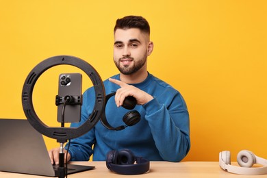 Photo of Technology blogger reviewing headphones and recording video with smartphone and ring lamp at wooden table on orange background