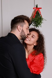 Happy man kissing his girlfriend under mistletoe bunch near light grey wall