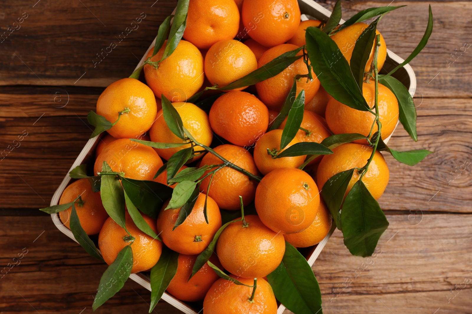 Photo of Fresh tangerines with green leaves in crate on wooden table, top view