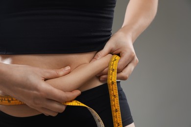 Photo of Woman with measuring tape touching belly fat on grey background, closeup. Overweight problem