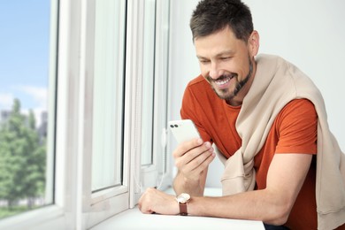 Photo of Happy man using smartphone near window, space for text