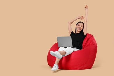 Photo of Cheerful woman with laptop sitting on beanbag chair against beige background, space for text