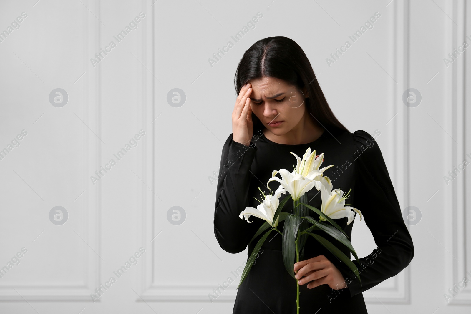 Photo of Sad woman with lilies mourning near white wall, space for text. Funeral ceremony