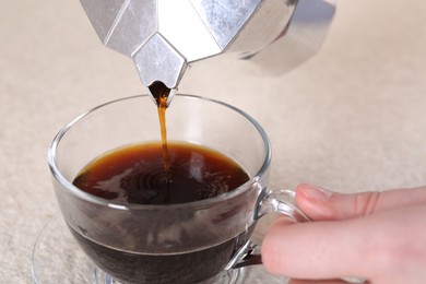 Woman pouring aromatic coffee from moka pot into glass cup at light textured table, closeup