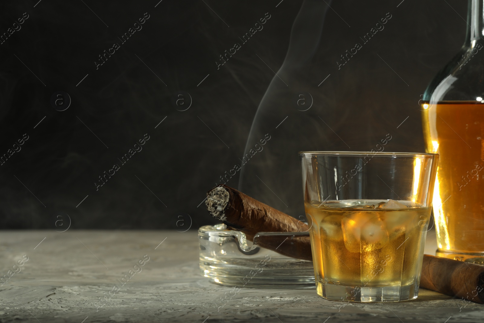 Photo of Bottle, glass of whiskey with ice cubes and cigars on grey table. Space for text