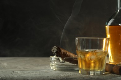 Bottle, glass of whiskey with ice cubes and cigars on grey table. Space for text