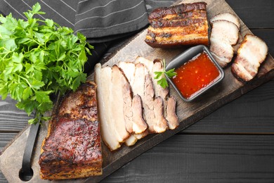 Photo of Pieces of baked pork belly served with sauce and parsley on black wooden table, top view