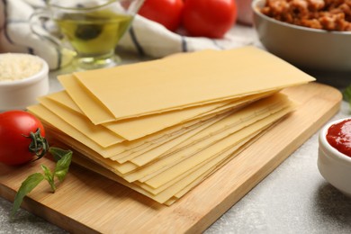 Photo of Cooking lasagna. Wooden board with pasta sheets and other products on grey textured table, closeup