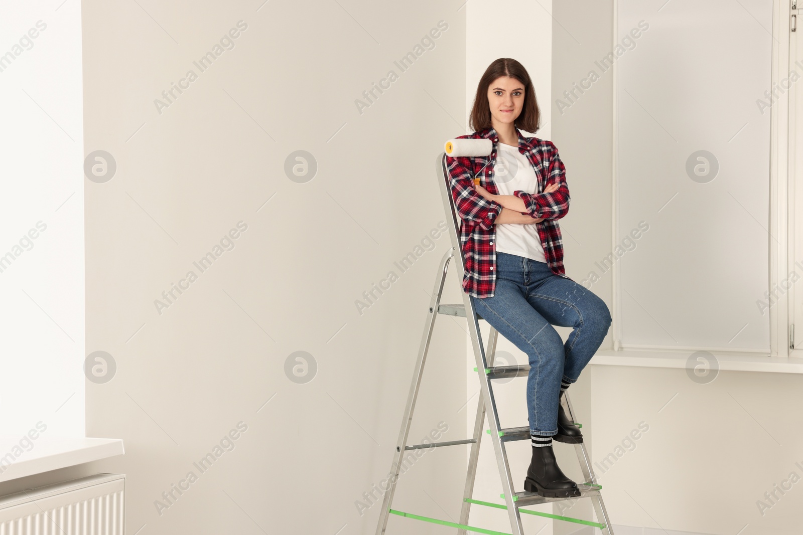 Photo of Young beautiful woman with roller on metal stepladder indoors. Room renovation