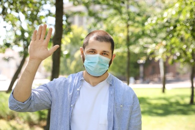 Photo of Man in protective face mask showing hello gesture outdoors. Keeping social distance during coronavirus pandemic