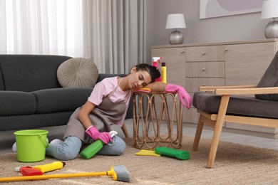 Tired young woman sitting on floor and cleaning supplies in living room