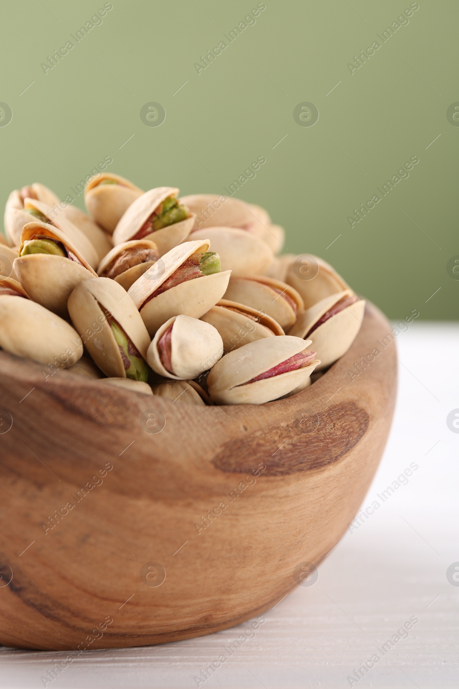 Photo of Tasty pistachios in bowl on white wooden table against olive background, closeup