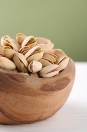 Tasty pistachios in bowl on white wooden table against olive background, closeup