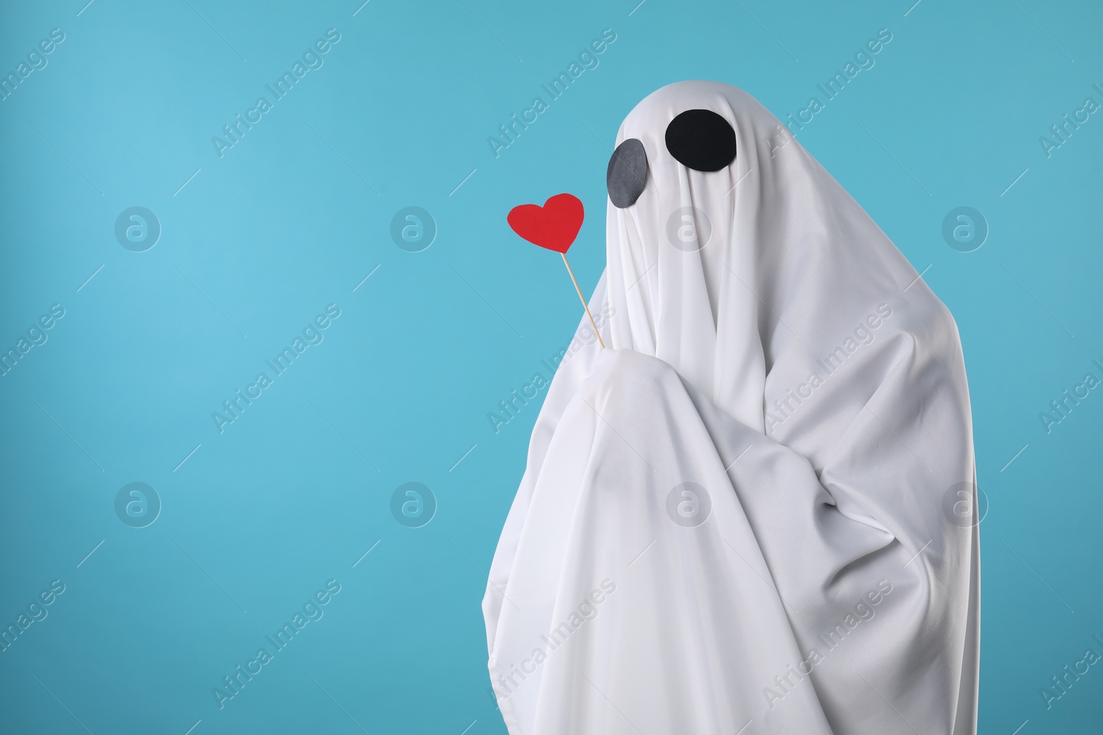 Photo of Cute ghost. Person covered with white sheet holding stick with red heart on light blue background, space for text